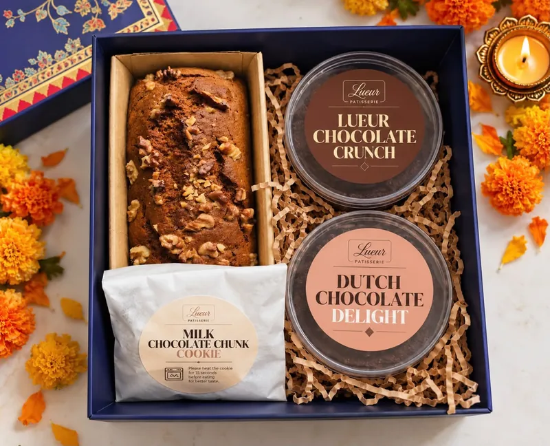 A navy gift hamper with a walnut loaf cake, a Milk Chocolate Chunk Cookie, Lueur Chocolate Crunch and Dutch Chocolate Delight, among marigolds and a diya