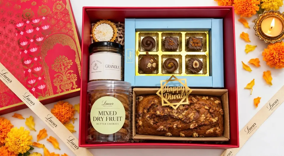 An open red Lueur gift hamper with a box of chocolates, a loaf cake with a Happy Diwali topper, granola and Mixed Dry Fruit butter cookies, among marigolds and a lit diya